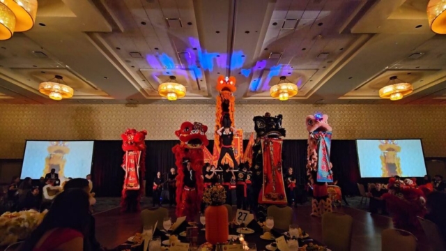 An indoor, wide eye-level shot capturing an acrobatic lion dance performance inside a large, dimly lit hotel ballroom with a high coffered ceiling and elegant chandeliers. In the center of the stage area, a performer stands balanced atop a high stack of tables, lifting another team member high overhead who operates the head of a tall orange ceremonial lion costume. Surrounding this central stunt, four other lions—in red, black, pink, and white—stand in a semi-circle displaying vertical festive banners. Large projection screens on the left and right mirror the live action, while round dinner tables with formal place settings are visible in the foreground.