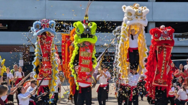 A vibrant outdoor performance during a Lunar New Year celebration featuring multiple colorful lion dance costumes surrounded by a burst of glittering gold confetti. In a dynamic row, four main lions are visible: a light blue and yellow lion on the left, a bright neon-green lion in the center-left, a white and yellow lion being lifted high by a performer in the center-right, and a bold red lion on the far right. The central green and rightmost red lions unfurl long red scrolls with decorative gold lettering, with the center scroll clearly reading "HAPPY NEW YEAR" and the right scroll reading "PROSPERITY". Performers dressed in white t-shirts, black pants, and red sashes animate the lions and hold up traditional festival flags in the background, while an appreciative crowd watches and takes photos from the sides under bright daylight.