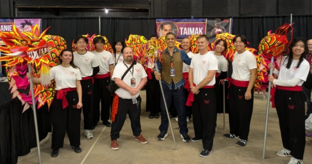 An indoor, wide eye-level group portrait capturing martial arts action star Taimak posing with members of the Texas Dragon/Lion Dance Team inside a convention hall. In the center, Taimak smiles while wearing a brown vest over a blue denim shirt, holding a silver pole topped with a decorative golden dragon pearl prop. He is surrounded by student team members wearing matching white t-shirts, black performance pants, and red sashes. On the far left, a female performer holds a pole supporting the large, vibrant red and yellow head of a ceremonial dragon costume, whose long body extends behind the group. In the background, black drapery and a convention banner reading "TAIMAK" are visible.