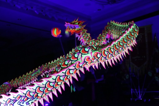An indoor, wide low-angle action shot capturing a traditional luminous dragon dance performed under dark blacklight conditions. The long, winding ceremonial costume is heavily decorated with glowing fluorescent neon colors, showcasing bright pink, yellow, and green scales and spikes that illuminate under the ultraviolet lighting. In the center background, the glowing head of the luminous dragon faces a small, glowing spherical dragon pearl prop held up on a pole. The human performers maneuvering the support poles are completely obscured by the dark, shadowy background of the indoor venue. Photo credit: ACU Residence Life.