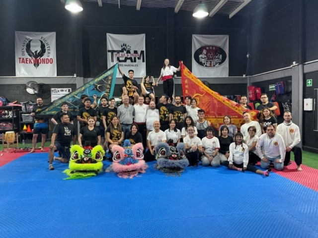 A large group portrait taken indoors on a blue and red interlocking martial arts floor mat inside a training facility. A diverse group of over thirty people stand, sit, and kneel together, representing an international collaboration. In the foreground on the floor rest three beautifully detailed Chinese lion dance heads in bright neon green, pink with blue accents, and dark blue with grey fur. Behind the group, several individuals hold up two massive triangular ceremonial flags—one teal and black with gold Chinese characters on the left, and one bright red and gold on the right. The back wall features various martial arts banners, including signs for Taekwondo and MMA, under industrial overhead lights.