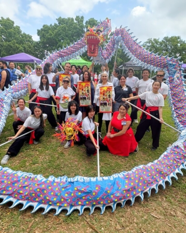A large outdoor group photograph taken on a grassy field at the Austin Dragon Boat Festival. Members of a dance troupe and local political representatives stand together, framed in a large circle by a long, undulating dragon dance costume held up on poles. The dragon features a detailed red and blue head at the top center, with a body patterned in bright blue, red, and yellow scales looping down to the foreground grass. In the center, Austin City Council District 6 Representative Krista Lane, Austin City Council District 7 Representative Mike Siegel, and Texas State Representative Lulu Flores stand alongside team members holding decorative red and gold ceremonial scrolls. The troupe members are dressed in matching white t-shirts, black pants, and red waist sashes, with some kneeling in the grass holding a colorful, fringed dragon ball prop under a bright sky with light clouds and green trees.