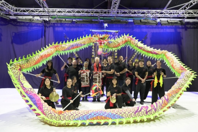 A group photograph on a stage featuring Austin Mayor Kirk Watson surrounded by members of a traditional dragon dance troupe. The troupe members are dressed in matching black t-shirts and black pants, with some wearing red sashes or holding traditional percussion instruments like cymbals and a gong. They hold up a long, vibrant dragon dance costume on poles, looping it completely around the group to frame everyone in a large circle. The dragon features a detailed red head at the top center and a body patterned with lime green, pink, yellow, and purple scales. In the center, Mayor Kirk Watson wears a festive red scarf and holds a traditional red and gold scroll alongside other members, posed against a dark blue background under bright stage lighting.