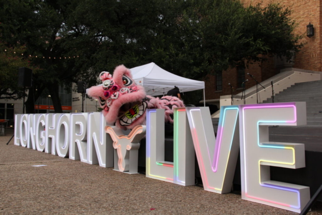 An outdoor shot of a pink and white traditional Chinese lion dance costume interacting with a large event display. In the foreground, large white marquee letters spell out "LONGHORN LIVE" on a gravel surface, with the word "LIVE" glowing with soft, colorful internal LED lights in shades of pink, green, yellow, and blue. Nestled between the two words is a matching white marquee cutout of the iconic University of Texas Longhorn silhouette, lit up with an orange border. Peeking out from behind the Longhorn symbol, a performer manipulates a vibrant pink lion head with large animated eyes, white fur accents, and a wide-open mouth showing stylized green and red details. In the background, a white canopy tent, lush green oak trees, and a brick campus building with outdoor concrete stairs are visible under an overcast sky.