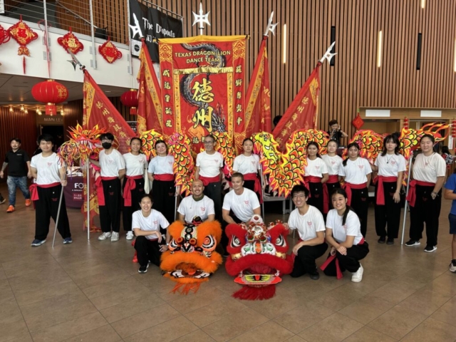 A large group portrait of the Texas Dragon and Lion Dance Team members gathered indoors in a spacious lobby with wooden slat wall paneling. In the center background stands a massive, ornate red banner reading "TEXAS DRAGON/LION DANCE TEAM" in gold letters, flanked by several traditional red flags on tall poles. Flanking the center banner, troupe members stand in a row holding up a long, colorful dragon dance costume featuring yellow, pink, and red scales. In the foreground, five team members kneel on the tiled floor around two beautifully detailed lion heads—one bright orange and black, and one vibrant red and white. All team members are dressed uniformly in white t-shirts with red logos, black pants, and red waist sashes, creating a cohesive team display beneath red hanging lanterns.