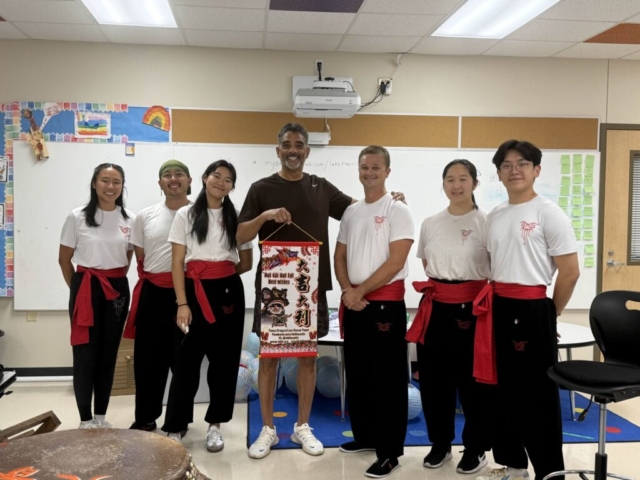 A group photograph taken inside a classroom or studio setting featuring six people standing in a row in front of a white whiteboard. Sifu Kisu stands in the center wearing a dark t-shirt and white sneakers, smiling as he holds up a decorative red and white hanging scroll that reads "Best Wishes" alongside Chinese characters and illustrations of a dragon and lion. Surrounding him are five members of a dance troupe dressed in matching white t-shirts with red logos, black pants, and traditional red sashes tied around their waists. A portion of a large performance drum is visible in the bottom-left foreground, and colorful classroom decorations, a projector, and a rolling office chair are visible in the background under bright indoor lighting.
