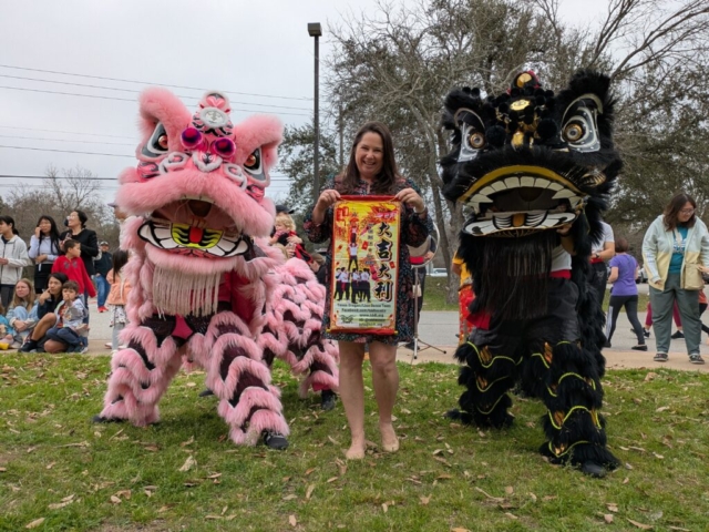 An outdoor, wide eye-level group portrait capturing Austin City Councilwoman Krista Laine posing with two ceremonial lion dance costumes from the Texas Dragon/Lion Dance Team on a grassy lawn. In the center, Laine smiles while holding up a decorative vertical red and yellow team poster displaying photos of an acrobatic stunt and official social media handles. Flanking her are two performers inside elaborate lion costumes: a pink and white Southern-style lion on the left and a black and gold lion on the right, both angled toward the center. In the background, a small crowd of local residents watches the library event under an overcast sky.