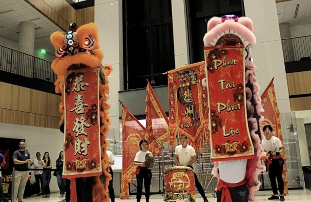 An indoor, wide eye-level shot capturing a traditional cultural performance inside a modern, multi-story building atrium with high ceilings and balconies. In the foreground, two standing ceremonial lion dance costumes present vertical red banners with decorative gold borders and text. On the left, an orange lion displays a banner written in Chinese calligraphy. On the right, a pink and white lion displays a banner reading "Phát Tài Phát Lộc" in Vietnamese. In the center background, members of the Texas Dragon/Lion Dance Team provide percussion accompaniment, featuring a female student striking a hand gong, a male student playing a large traditional drum, and a third student holding cymbals. A small group of spectators watches from the lower left.
